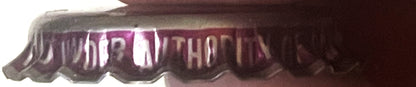 Close-up of a ridged metal bottle cap with purple and white striped design and embossed text, shown at an angle against a dar