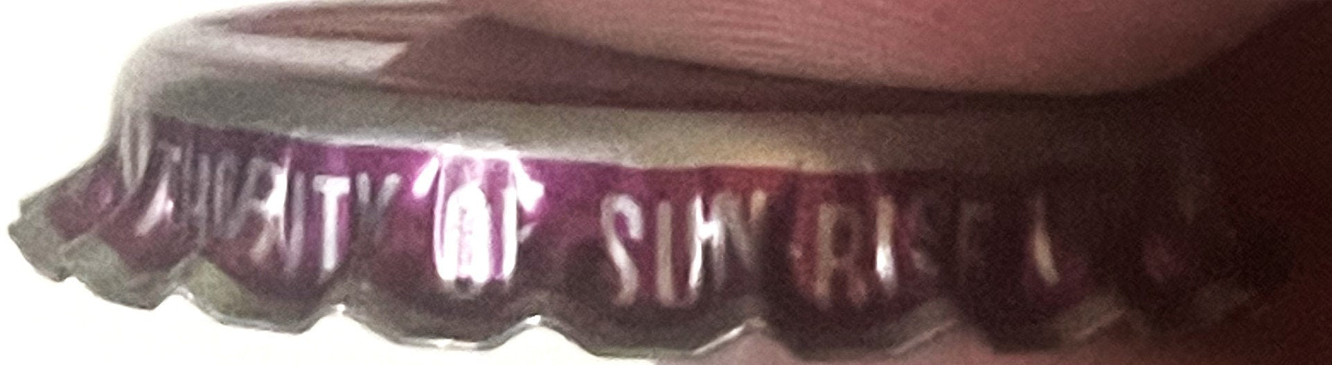 Close-up of vintage bottle cap with crimped burgundy and white ridged edges, white text reading "SUN RISE" visible on dark ba