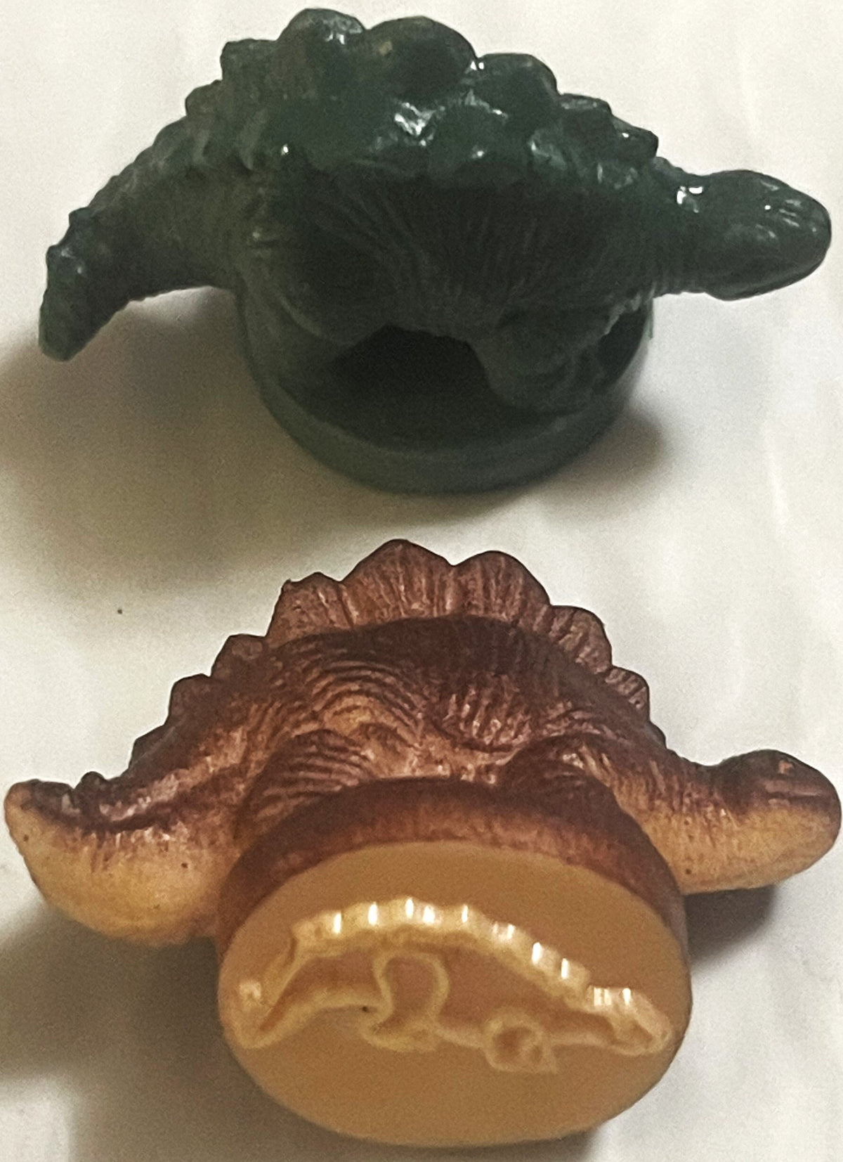 Two vintage rubber dinosaur stamps: a dark green armored dinosaur and a brown plate-backed dinosaur turned to show the raised dinosaur outline on its base