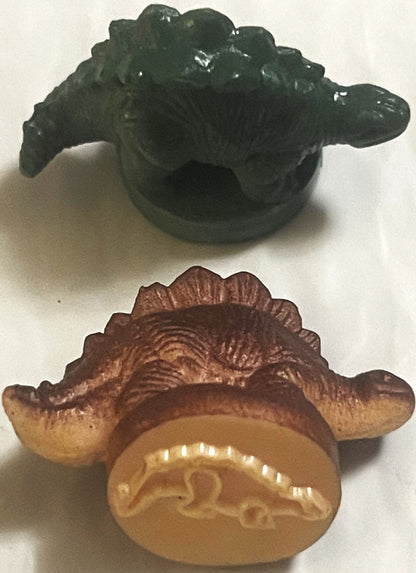 Two vintage rubber dinosaur stamps: a dark green armored dinosaur and a brown plate-backed dinosaur turned to show the raised dinosaur outline on its base