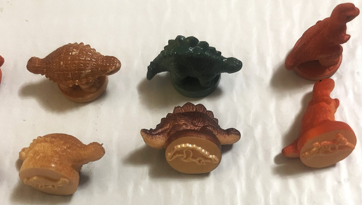 Six colorful rubber dinosaur stamps in brown, tan, orange, and dark green arranged on white background, showing embossed