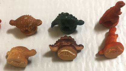 Six colorful rubber dinosaur stamps in brown, tan, orange, and dark green arranged on white background, showing embossed