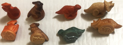 Eight small colored rubber dinosaur and animal stamps arranged on white surface, showing red, brown, tan, and green vintage t