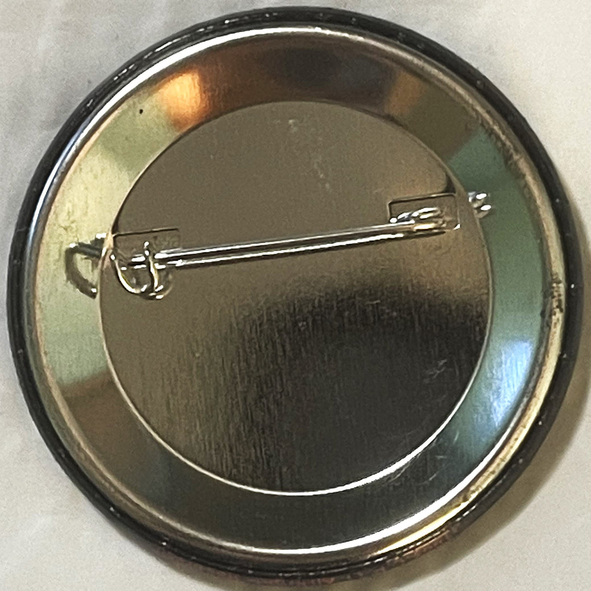 Back of metal pin button showing silver clasp against dark brushed surface with colored rim reflections