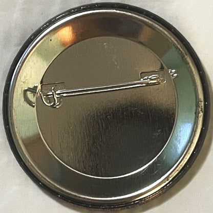 Back of metal pin button showing silver clasp against dark brushed surface with colored rim reflections