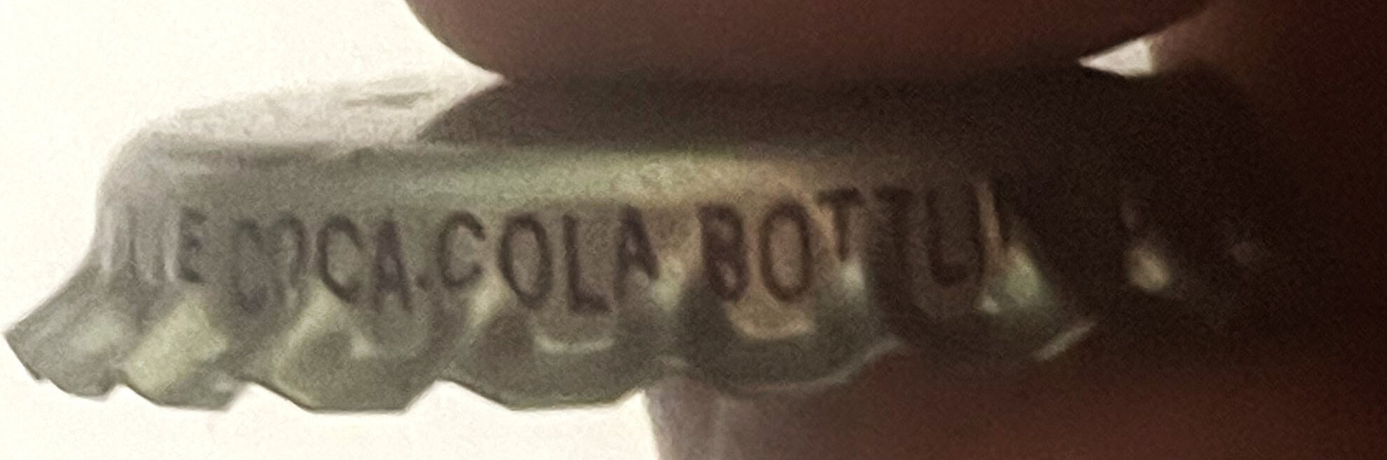 Close-up of vintage bottle cap showing embossed text "COCA COLA BOTTL" and partial branding, held against dark background for
