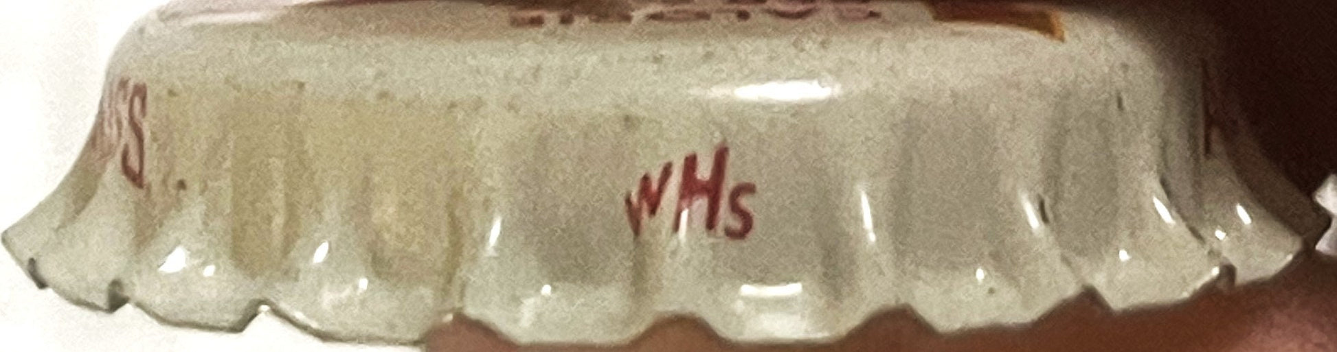 Cream-colored vintage bottle cap with scalloped edges and red "WHS" text printed on top, photographed from above
