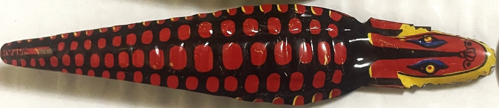 Red and black tin snake toy with yellow head, gold eyes, and mouth mechanism, viewed from above against white background