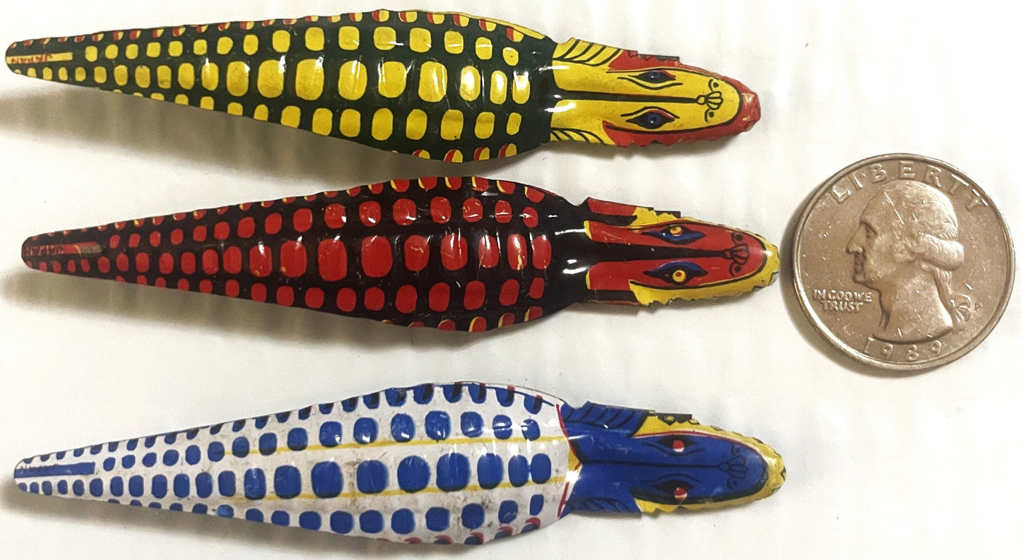 Three colorful vintage tin snake clickers in yellow-black, red-black, and blue-white with detailed heads, displayed next to a