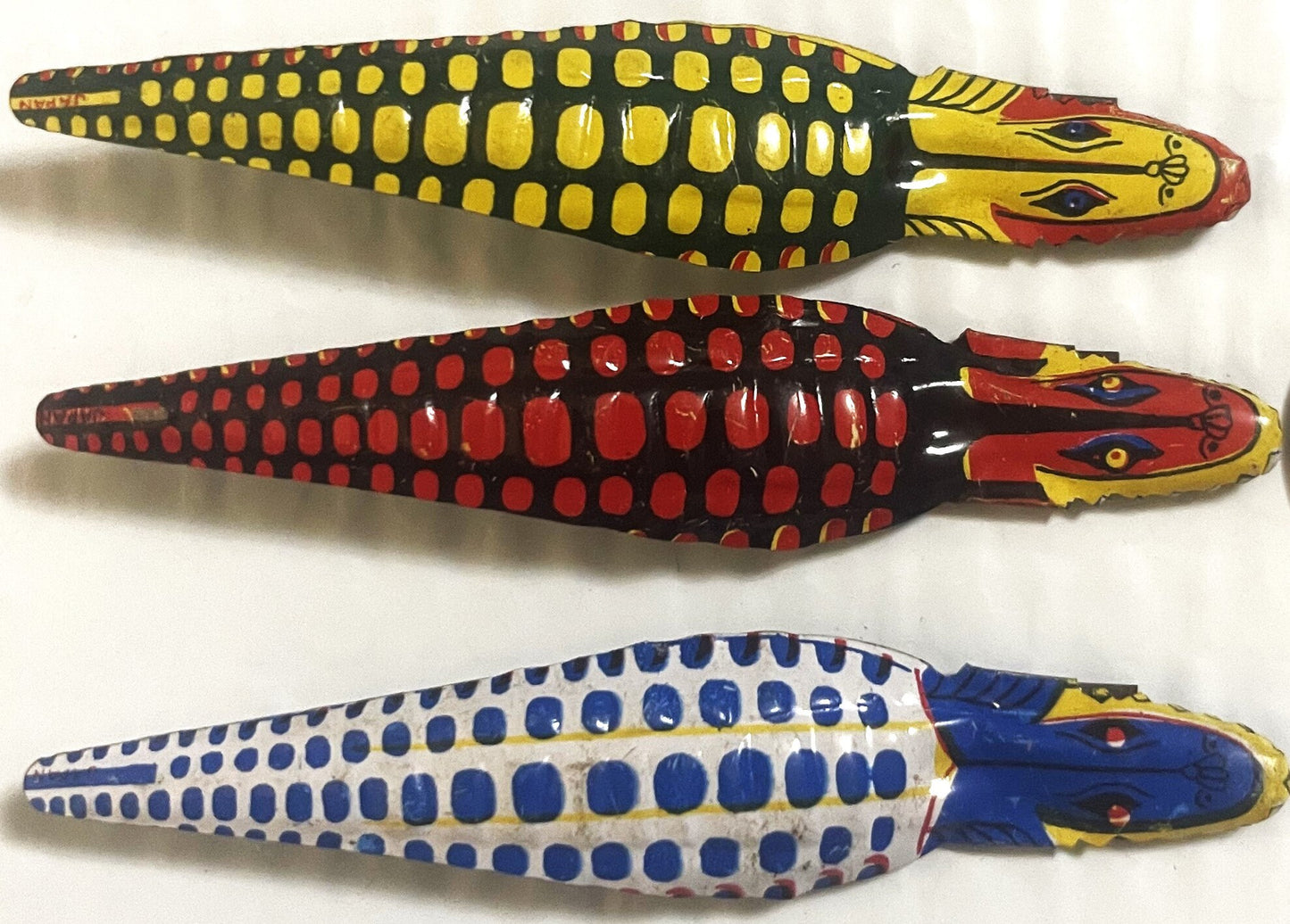 Three vintage tin snake clickers in yellow/black, red/black, and blue/white with articulated jaws, displayed against a white 
