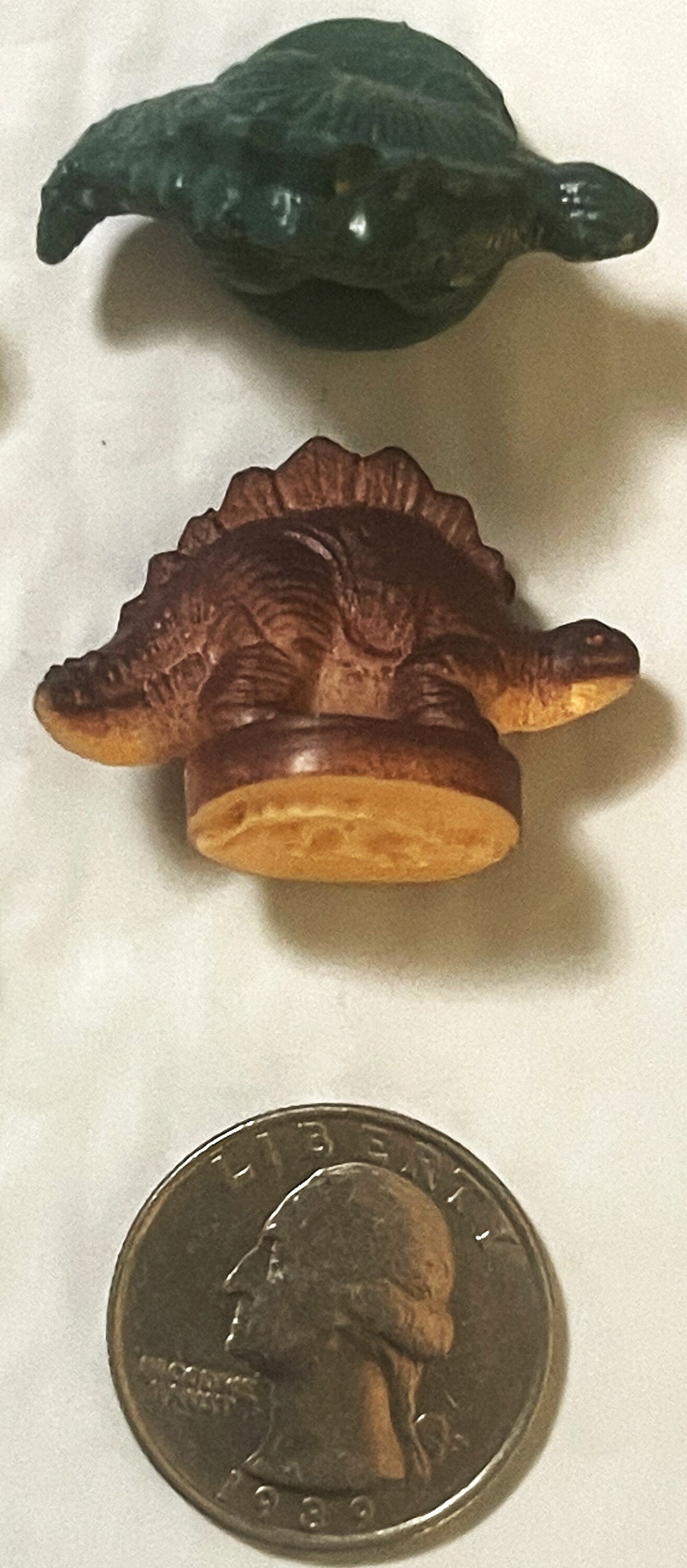 Two vintage rubber dinosaur stamps above a 1989 quarter for scale: a dark green armored dinosaur and a brown plate-backed dinosaur on its round stamp base