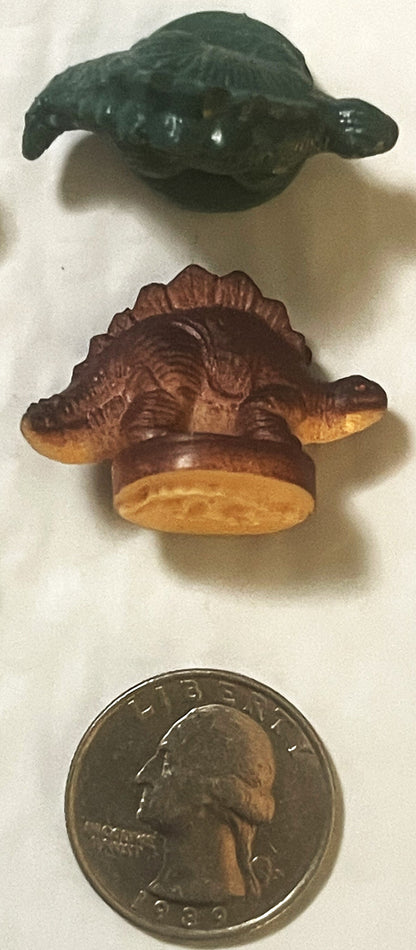 Two vintage rubber dinosaur stamps above a 1989 quarter for scale: a dark green armored dinosaur and a brown plate-backed dinosaur on its round stamp base