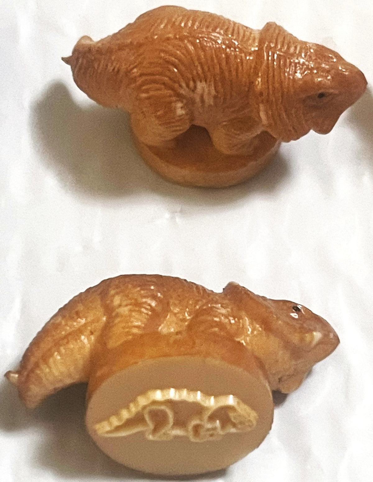Two tan rubber dinosaur stamps shown from top and bottom angles, displaying sculpted texture and the raised dinosaur outline on the stamp base