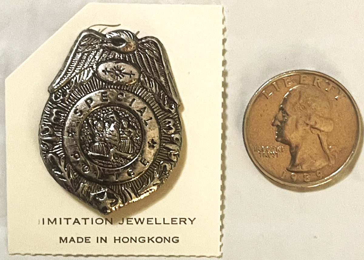 Silver police badge pin with eagle and shield design next to US quarter for scale, mounted on card labeled "Imitation Jewelle
