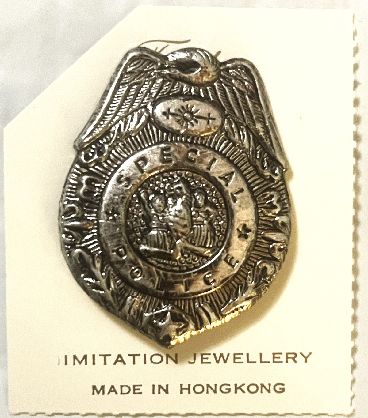 Silver-toned police badge pin with eagle on cream cardboard backing labeled "IMITATION JEWELLERY MADE IN HONGKONG