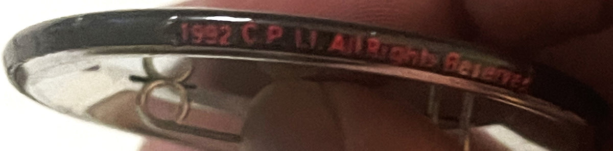 Close-up of vintage pin back button rim with red text and embossed design, held at angle showing metal edge and partial logo 