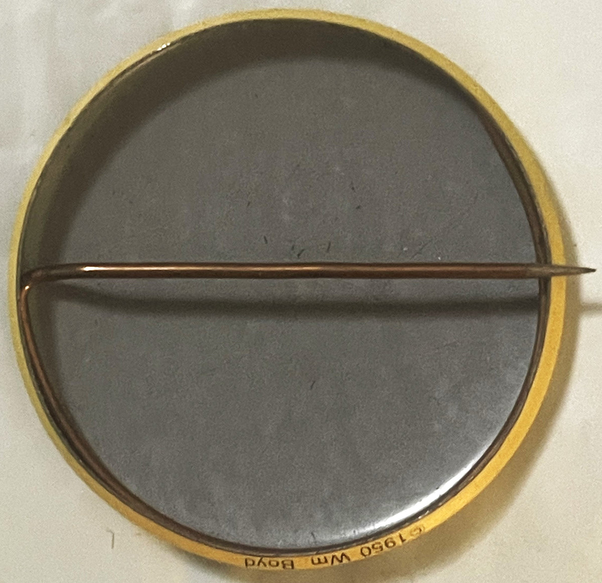Circular brass pin with gray enamel center divided by horizontal bar, text visible on rim reading "Quality Dairy St Louis