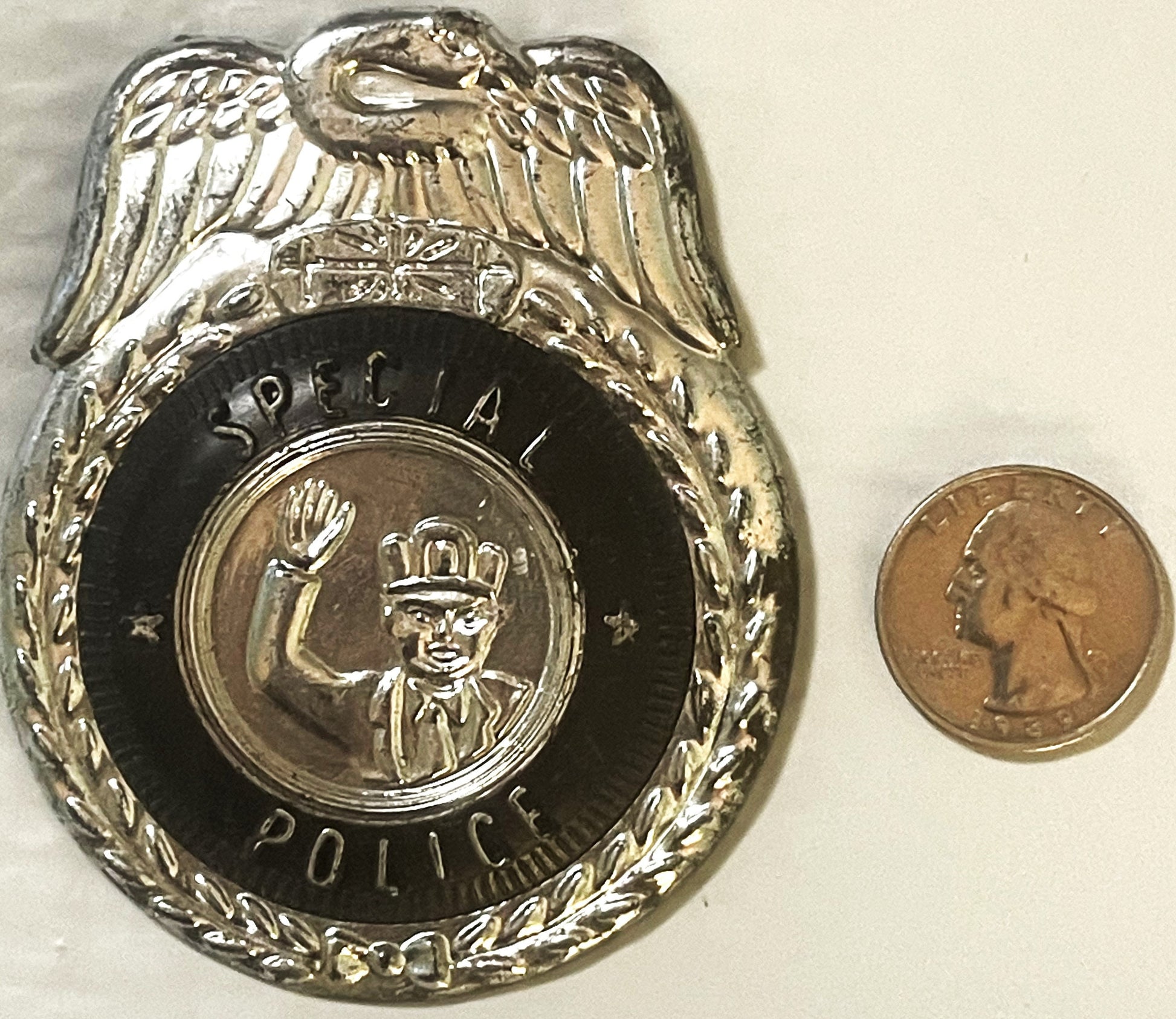 Silvered tin police badge with eagle on top, black enamel center showing crowned figure, next to quarter coin for scale