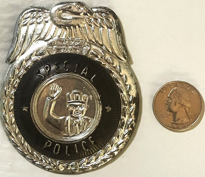 Silvered tin police badge with eagle on top, black enamel center showing crowned figure, next to quarter coin for scale
