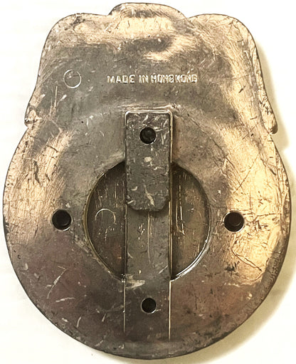 Back of vintage tin badge with "MADE IN HONG KONG" text, showing mounting holes and worn metallic surface
