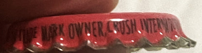 Close-up of red and green cork-lined crown bottle cap with embossed "Strawberry Crush" text, held by a thumb for scale