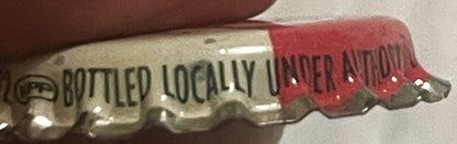 Close-up of vintage bottle cap showing "BOTTLED LOCALLY" text on cream and red crown with cork lining, held by finger for sca