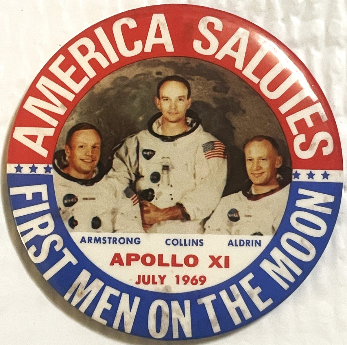 Front of the large 3.5 inch Apollo 11 pinback: the NASA crew portrait of Armstrong, Collins and Aldrin against a lunar backdrop, ringed by AMERICA SALUTES above and FIRST MEN ON THE MOON below, with APOLLO XI and JULY 1969 beneath the photograph.