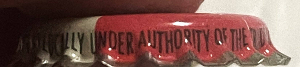 Close-up of red and silver cork-lined crown cap with "UNDER AUTHORITY OF THE" text visible on ridged metal surface
