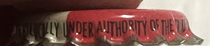 Close-up of red and silver cork-lined crown cap with "UNDER AUTHORITY OF THE" text visible on ridged metal surface