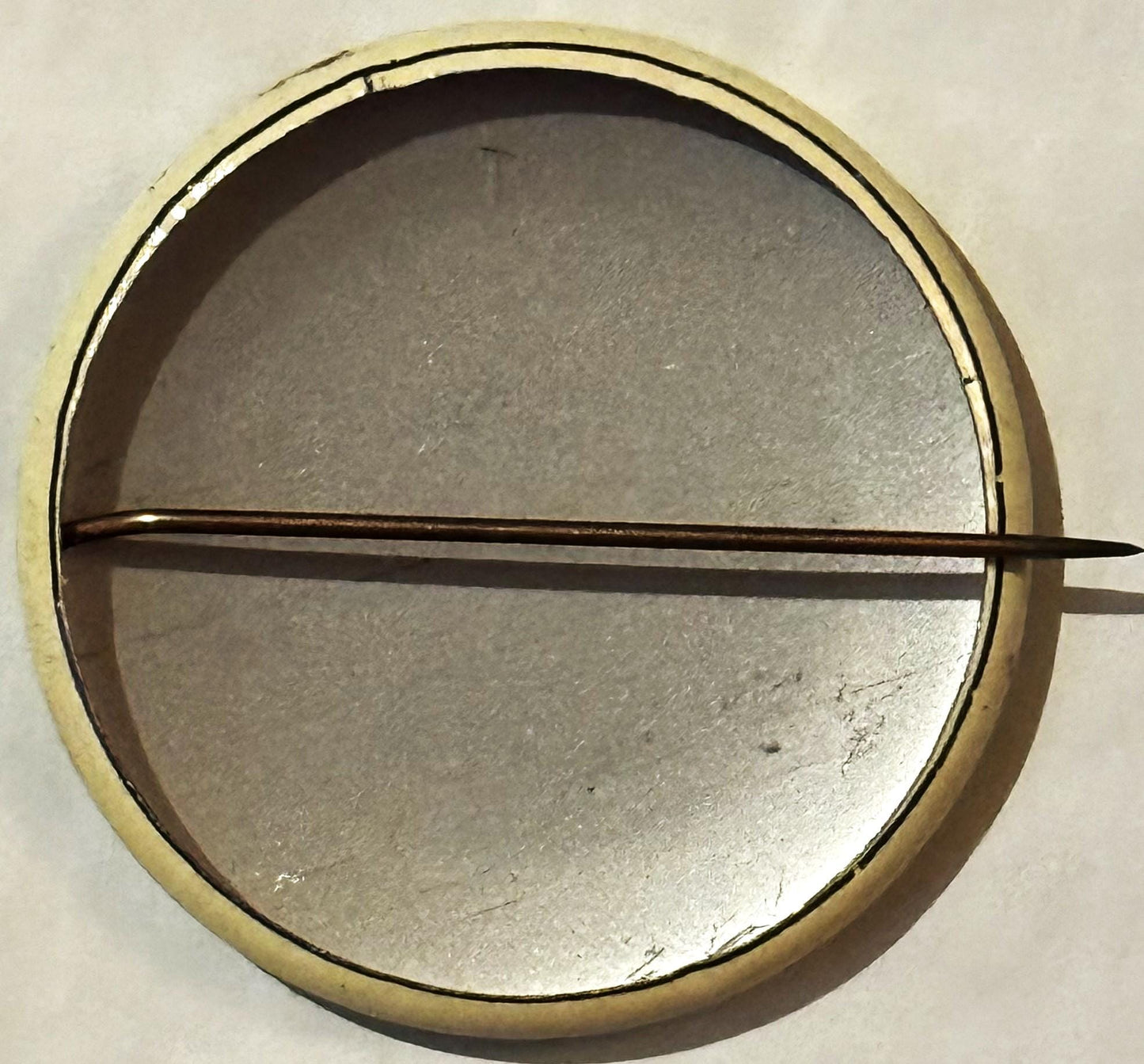 Circular metal pinback button with cream-colored rim and metallic center, divided horizontally by a dark pointer or needle, s