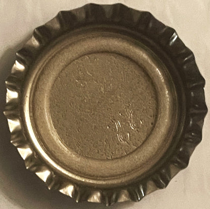 Unused metal bottle cap with crimped ridged edges shown flat against beige background, displaying worn tan surface with faint