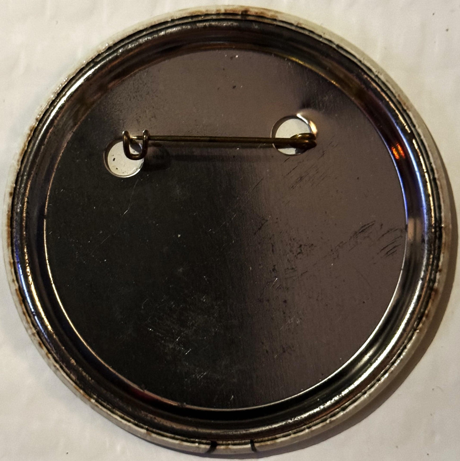 Back of circular pinback button with metal clasp and pin against white background, dark surface visible