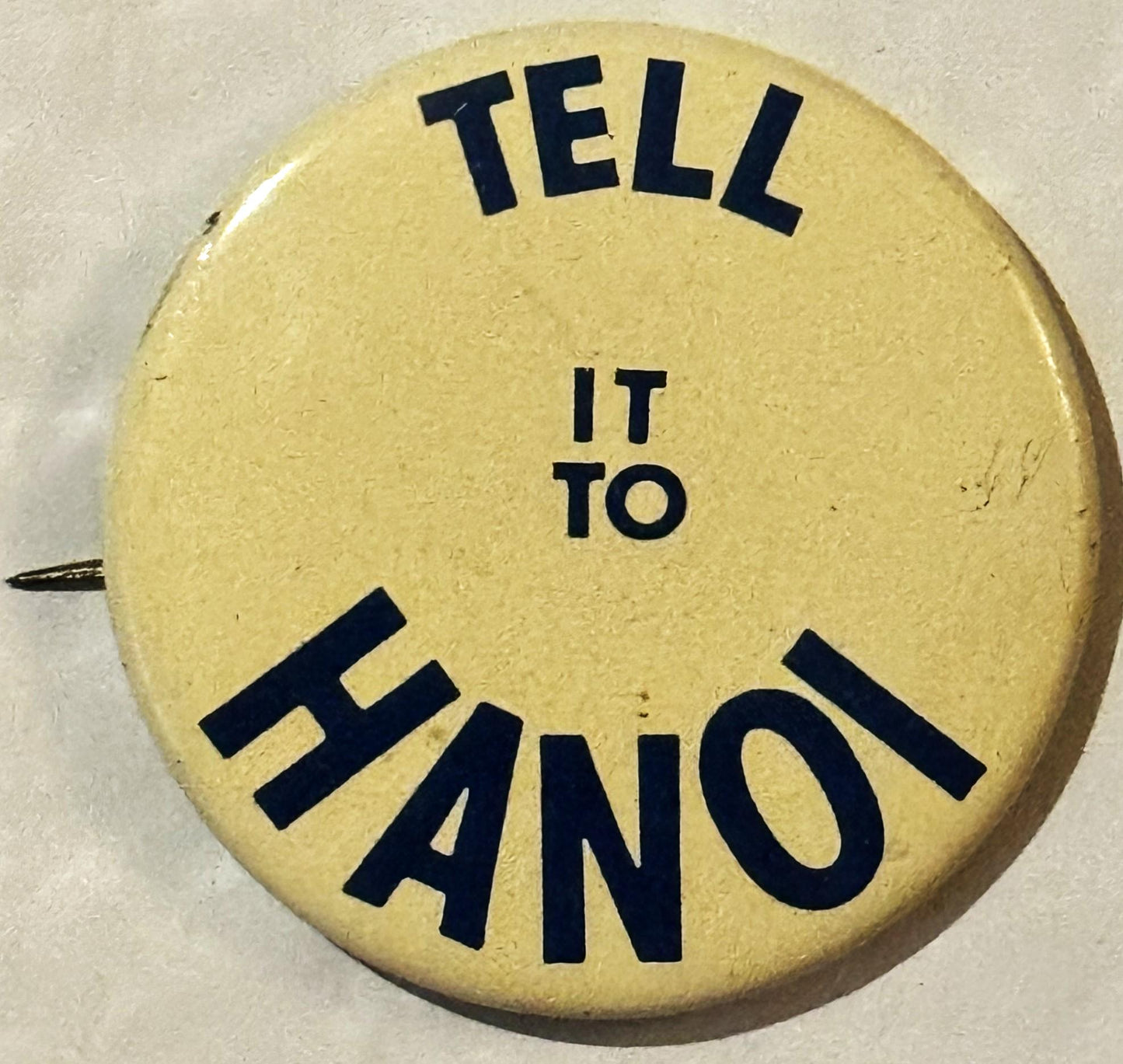 Circular cream-colored pinback button with black text reading "TELL IT TO HANOI" arranged around the edge, shown against beig