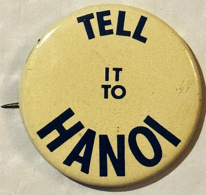 Circular cream-colored pinback button with black text reading "TELL IT TO HANOI" arranged around the edge, shown against beig