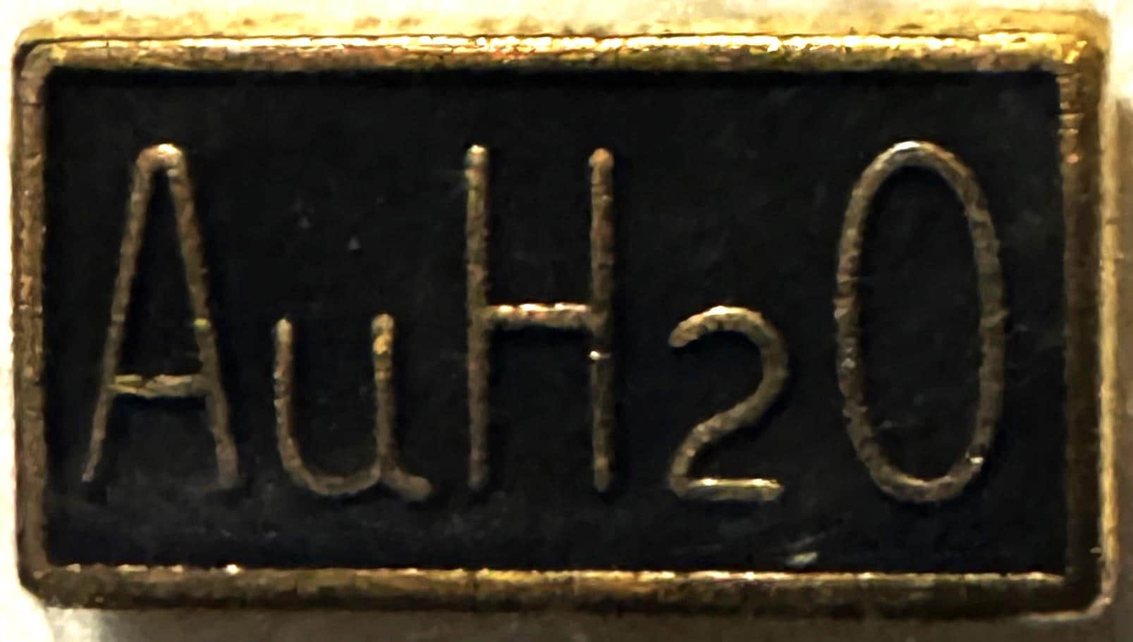 Close-up of embossed metal campaign pin showing "AuH2O" text on black background with gold-toned border and raised lettering