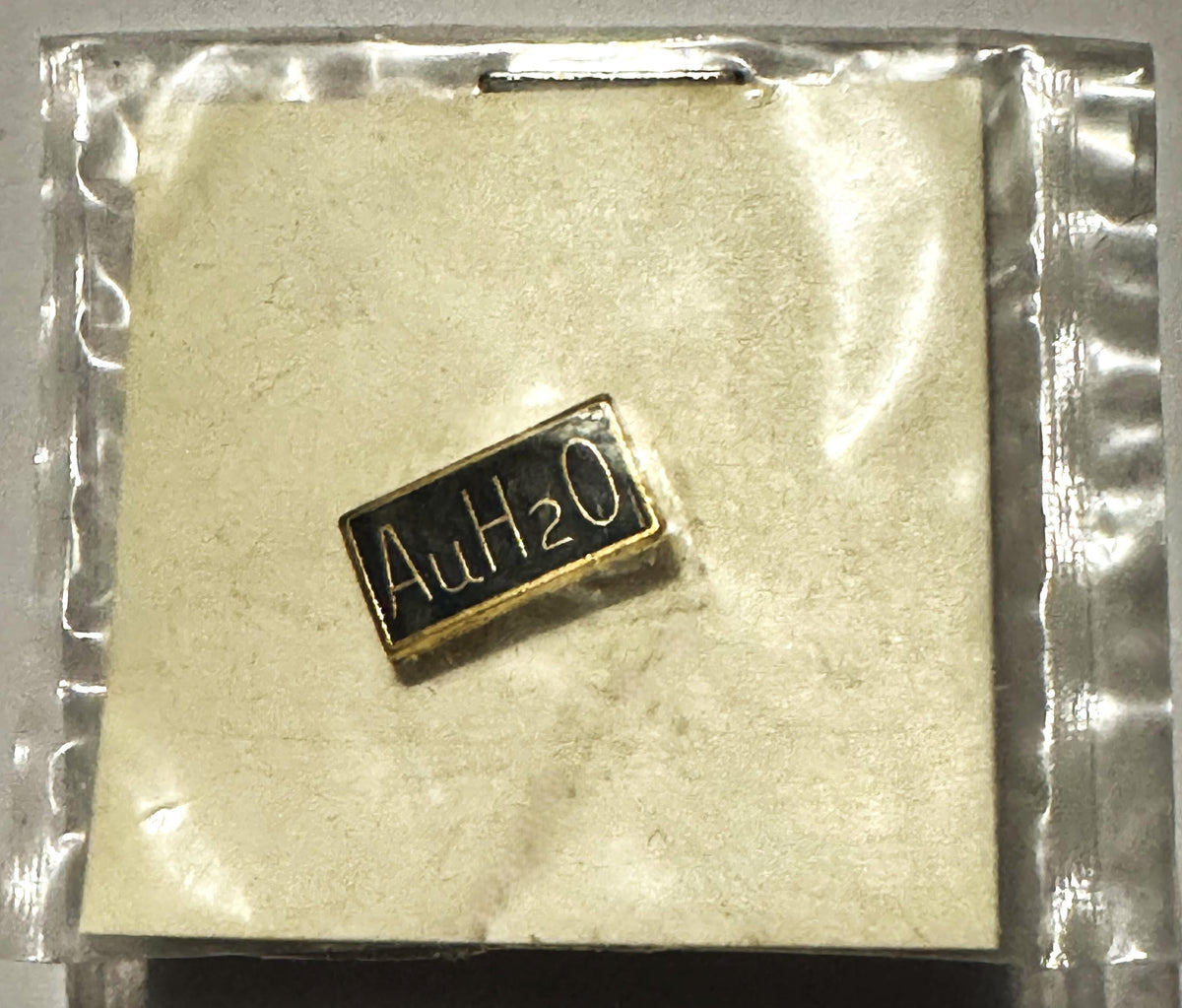 Small gold and black rectangular pin reading "AuH2O" on cream-colored backing in clear plastic sealed package