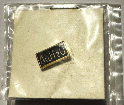 Small gold and black rectangular pin reading "AuH2O" on cream-colored backing in clear plastic sealed package
