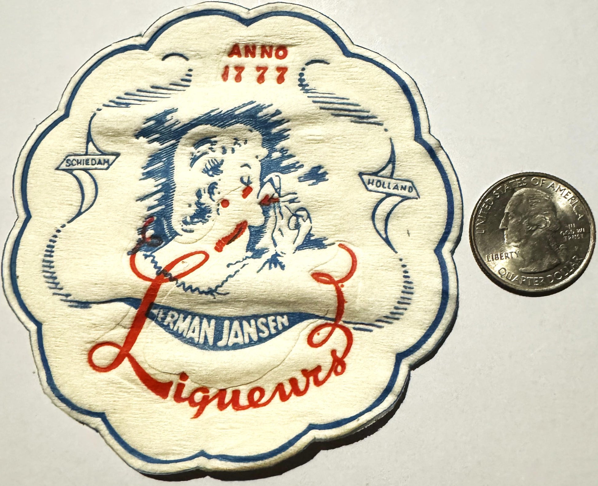 Vintage cloth coaster with Dutch lady drinking liqueur, blue and red details, dated 1777, next to quarter for scale.