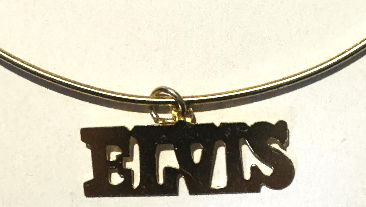 Black metal "ELVIS" nameplate charm hanging from gold-tone choker necklace against cream background