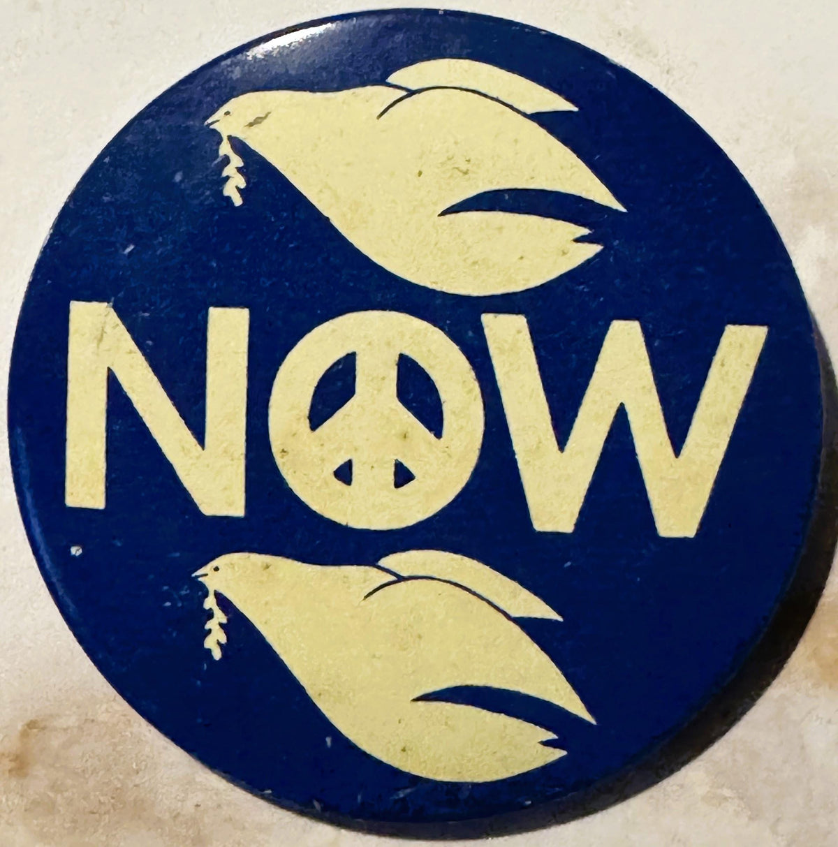 Vintage circular pinback button with navy blue background, cream-colored text reading "NOW" with peace symbol, flanked by two