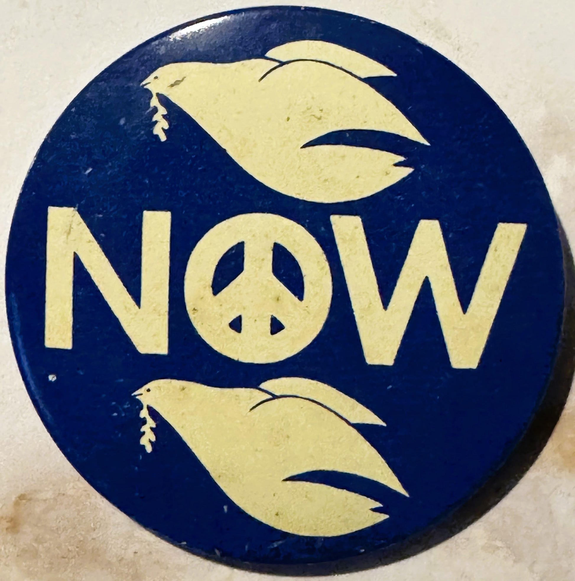 Circular navy blue button with yellow text reading "NOW" and a peace symbol, flanked by two yellow doves on cream background