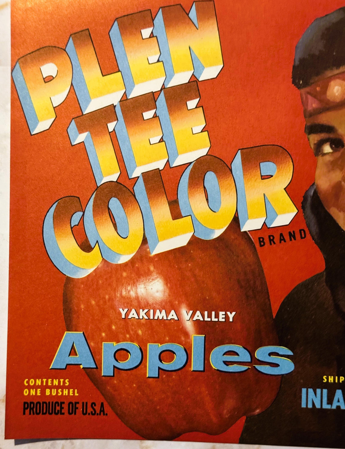 Vintage Plen Tee Color Brand apple crate label featuring bold 3D lettering and a large red apple on orange background, held b