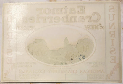 Cream-colored vintage cranberry barrel label with embossed oval landscape vignette showing sunrise over water and buildings, 