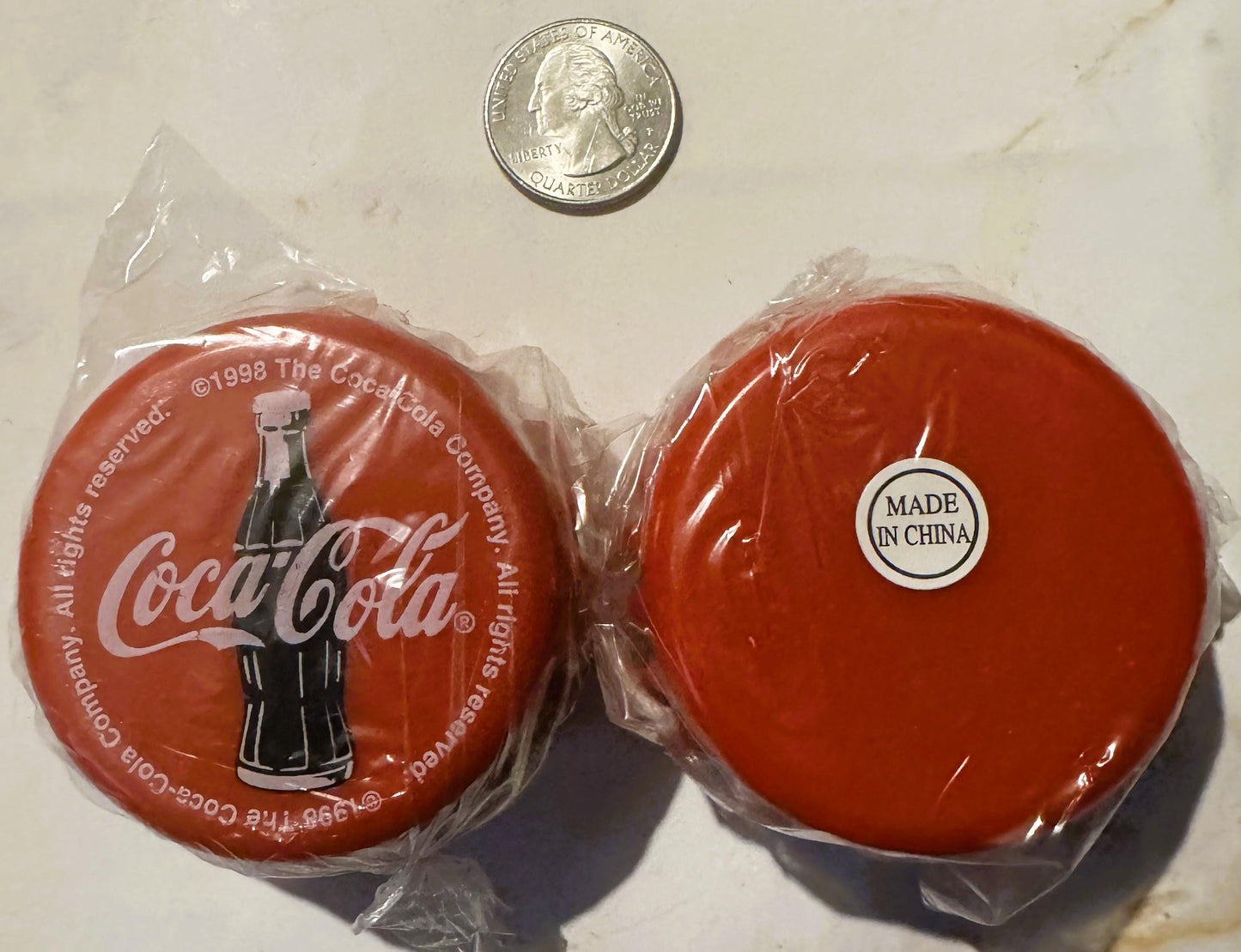 Two red Coca-Cola yo-yos in plastic packaging with a quarter for scale, one showing the bottle logo and 1998 copyright, the o