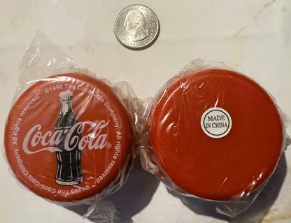 Two red Coca-Cola yo-yos in plastic packaging with a quarter for scale, one showing the bottle logo and 1998 copyright, the o