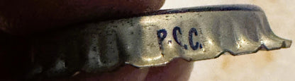 Vintage cream soda bottle cap with "P.C.C." embossed text, held in hand showing cork lining and weathered metal surface