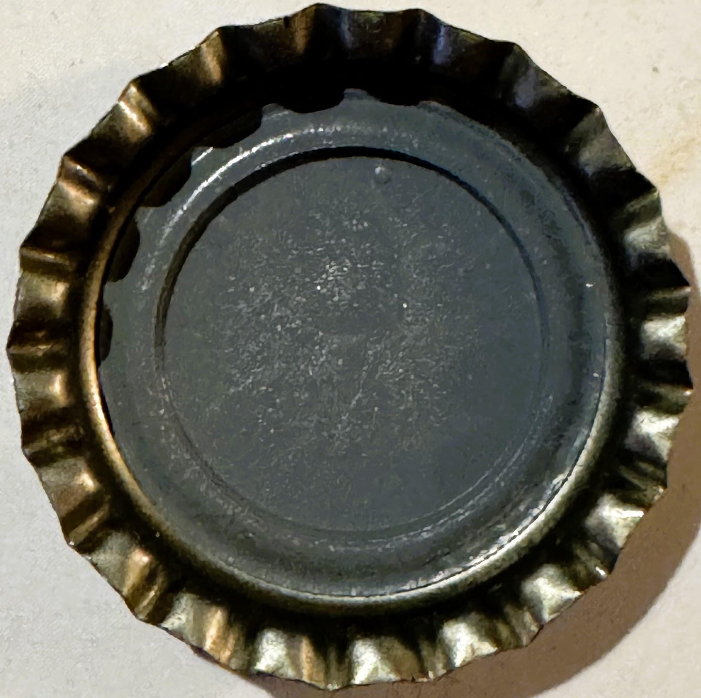 Vintage metal bottle cap with crimped gold-colored rim and dark center, photographed flat from above on white background