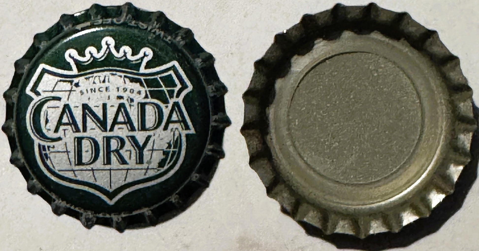 Front and back of a green and white Canada Dry bottle cap from 1904, showing crown logo and embossed globe design with blank 