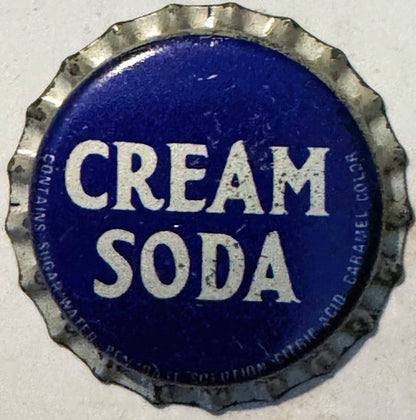 Vintage metal bottle cap with ridged edge, deep blue center, white lettering reading "CREAM SODA" with partial text visible a