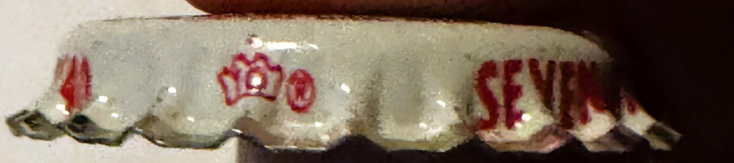 Vintage bottle cap with cream and red coloring, showing crown design and partial text reading "SEVEN" and other faded letteri